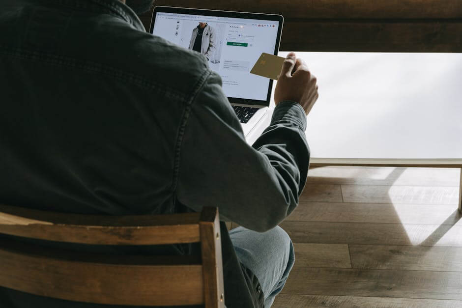 Adult man using a laptop while holding a credit card for online shopping in a comfortable indoor setting.